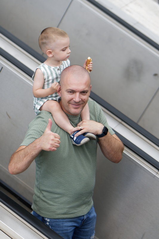 22 - Man wearing a green crewneck t-shirt with a textured fabric, carrying a little child, while giving a thumbs-up gesture